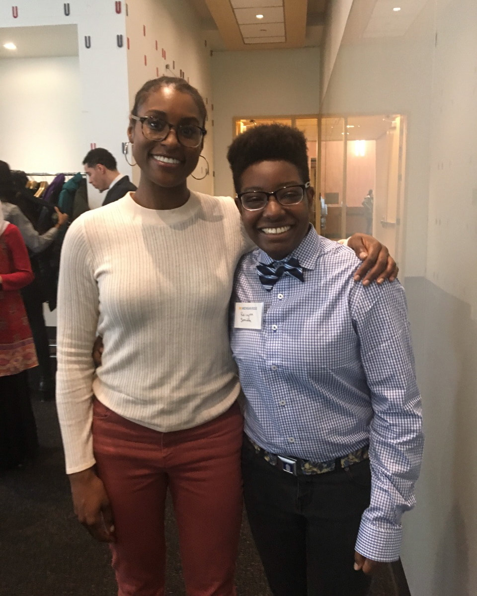 Raivynn, wearing blue plaid button up and blue bow tie, smiles next to a smiling Issa Rae, wearing yellow sweater.